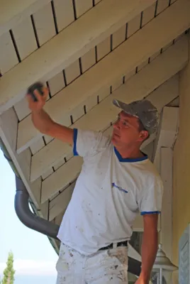 Le bras tendu vers le haut, l’artisan peint les lames sous le toit